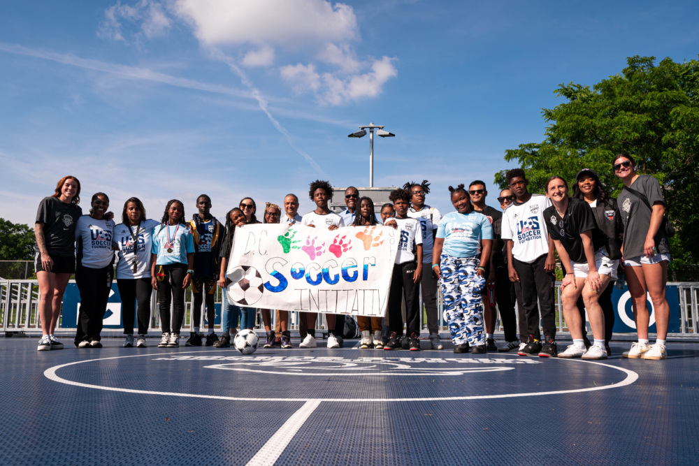 youth participants, adults, and women athletes pose for a photo at the center circle of the blue mini-pitch. they are also holding a white banner decorated with the text D.C. Soccer Initiative in big letters across the middle.